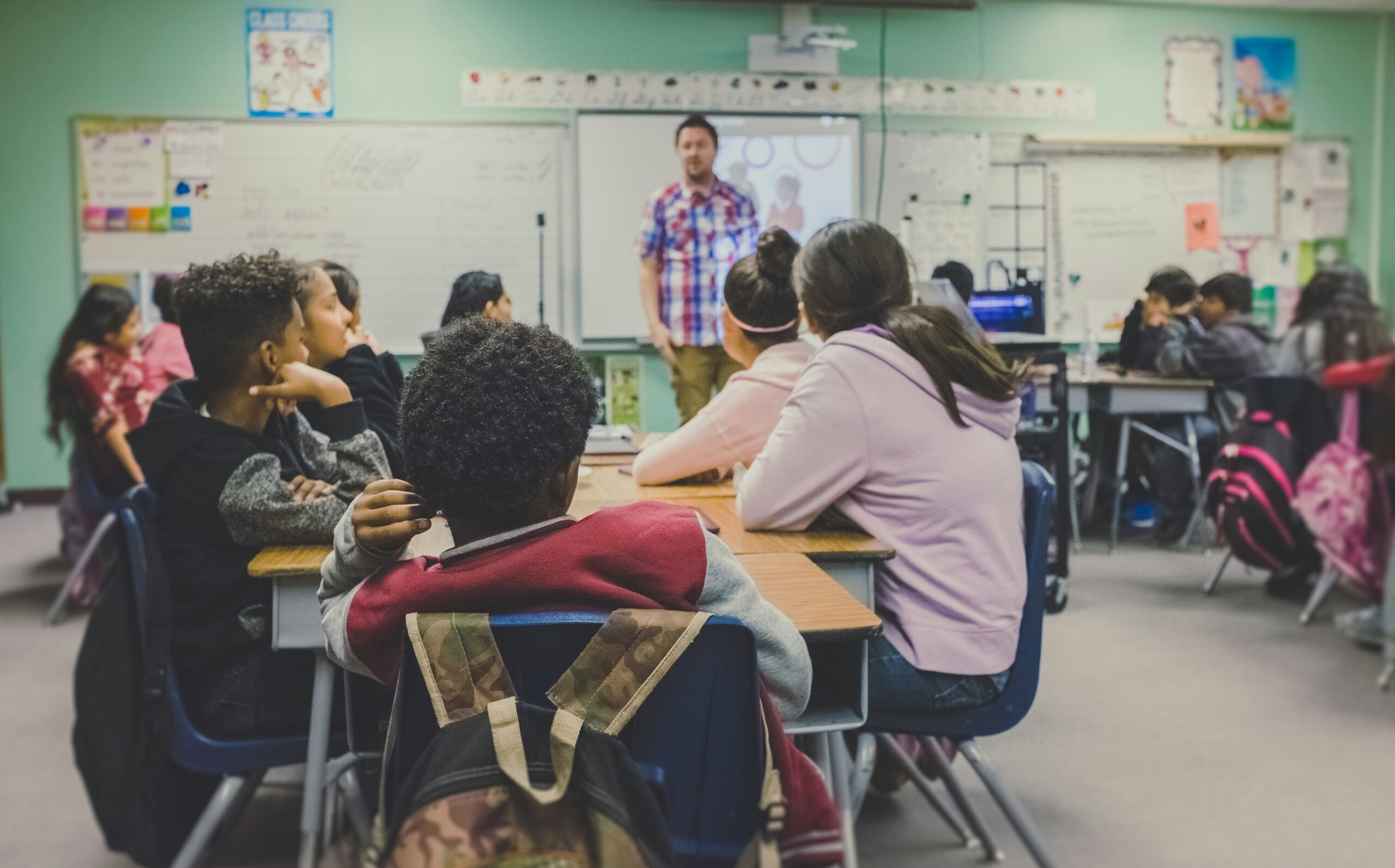 Students in a classroom listen to a teacher giving a lesson at the front.